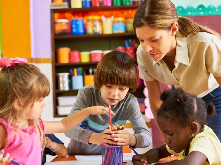 Drei Grundschüler und eine Lehrerin räumen Buntstifte in einen Becher.