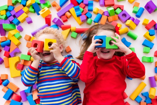Zwei Kinder liegen zwischen Bausteinen und gucken durch welche hindurch.