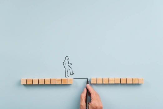 Eine Hand zeichnet mit einem Stift eine Linie über einen symbolischen Abgrund, den nun eine gezeichnete Silhouette überwinden kann. 